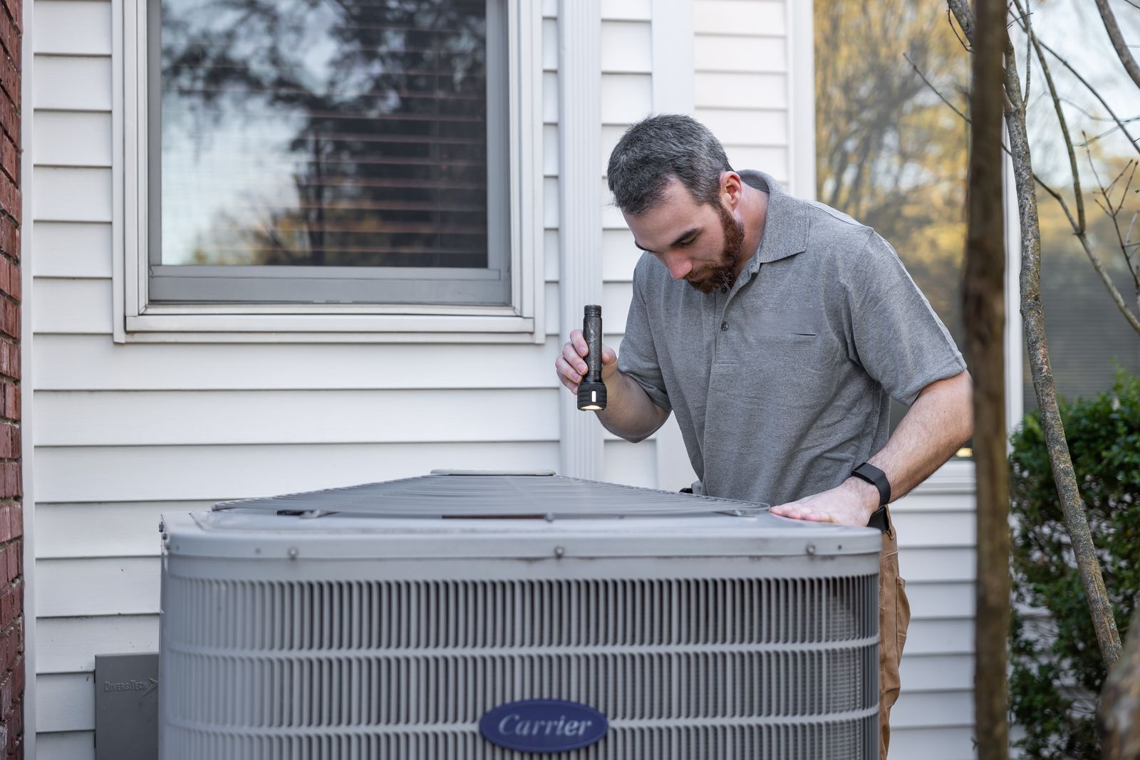 HVAC technician using a flashlight to inspect an outdoor air conditioning unit beside a home